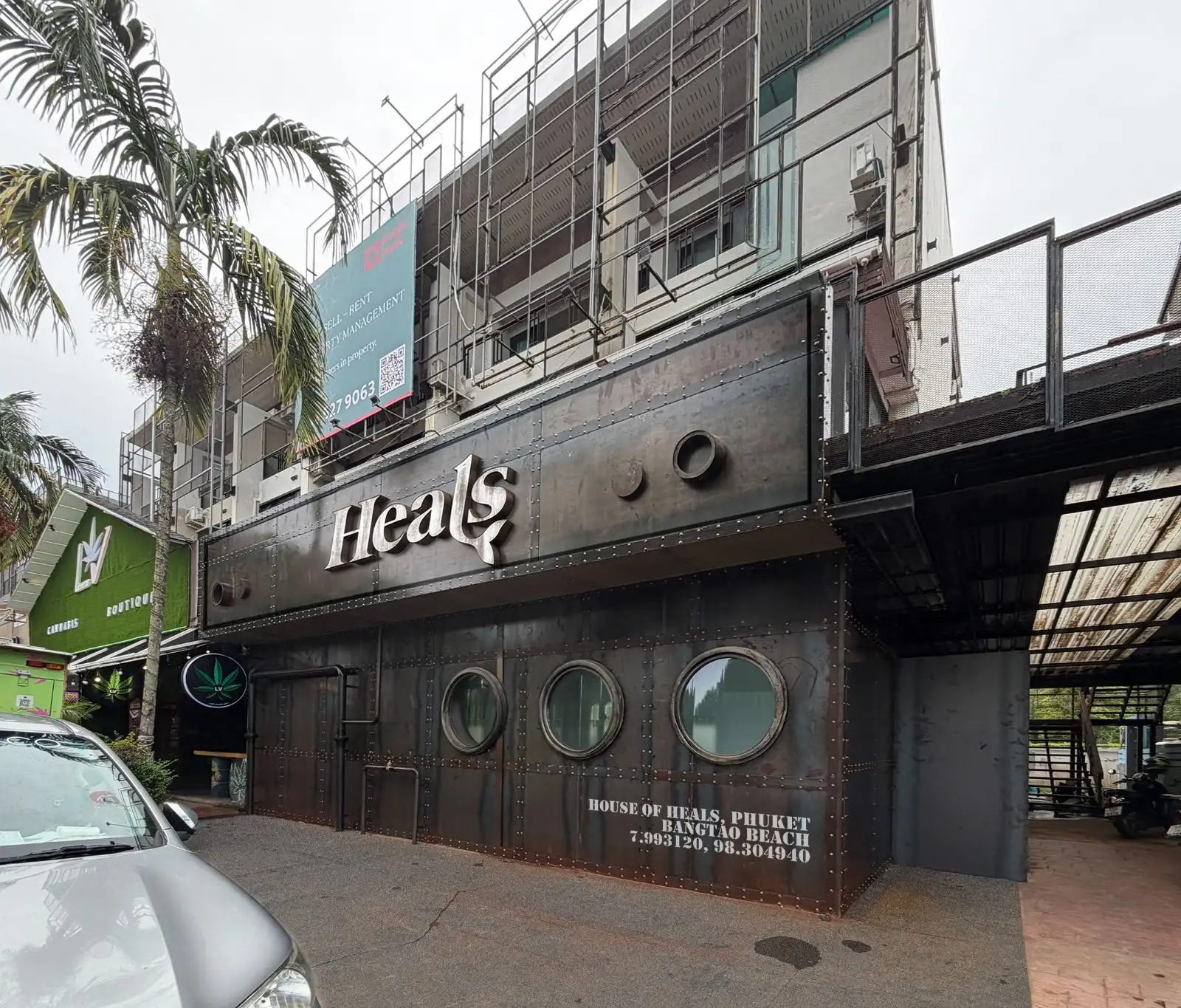 The Heals Phuket façade by day: dark riveted steel, three portholes and chrome Heals letters